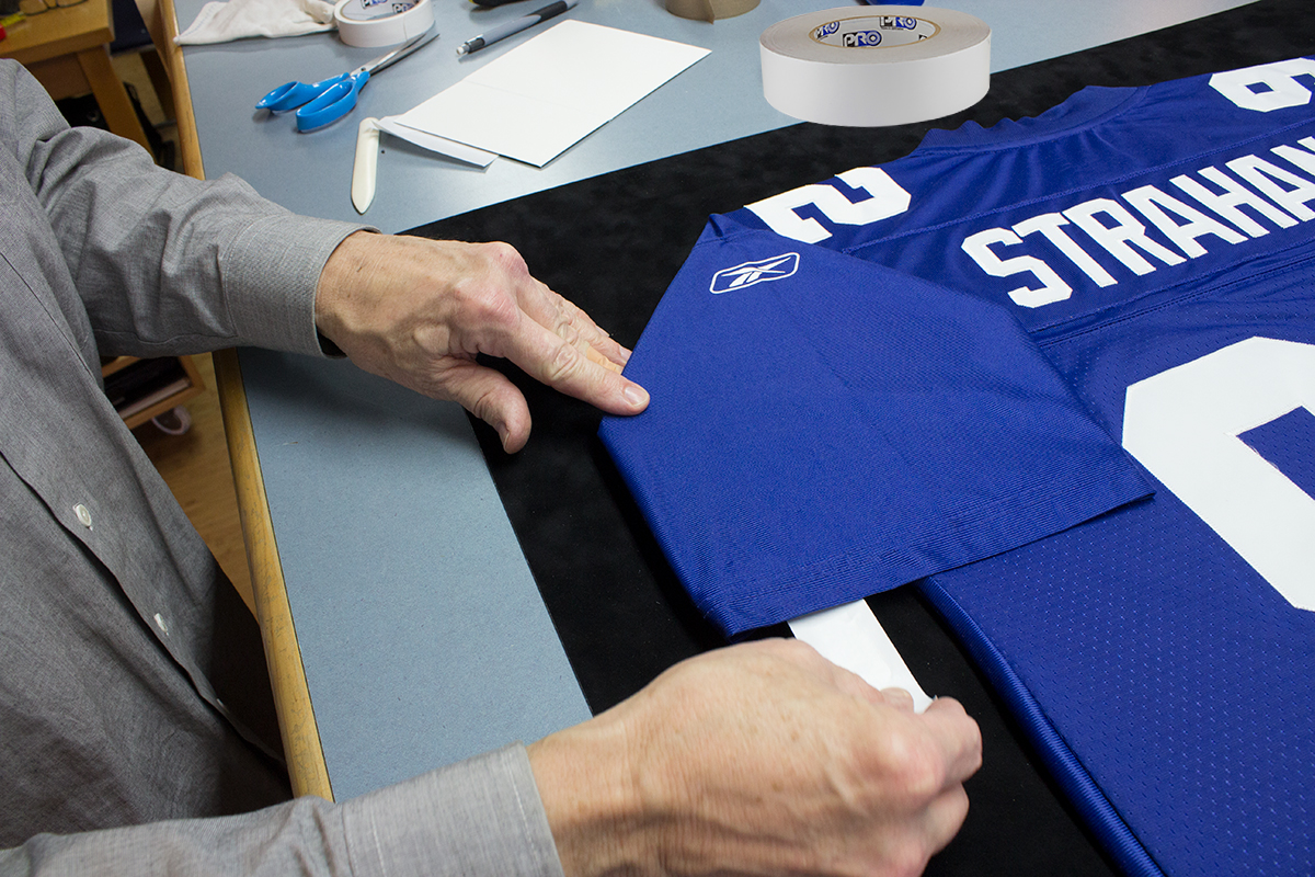 Our framer shown using Pro® Stitchery Tape to secure the sleeve of a jersey as he removes the tape liner to make the final bond. Our framer shown using Pro® Stitchery Tape to secure the sleeve of a jersey as he removes the tape liner to make the final bond.